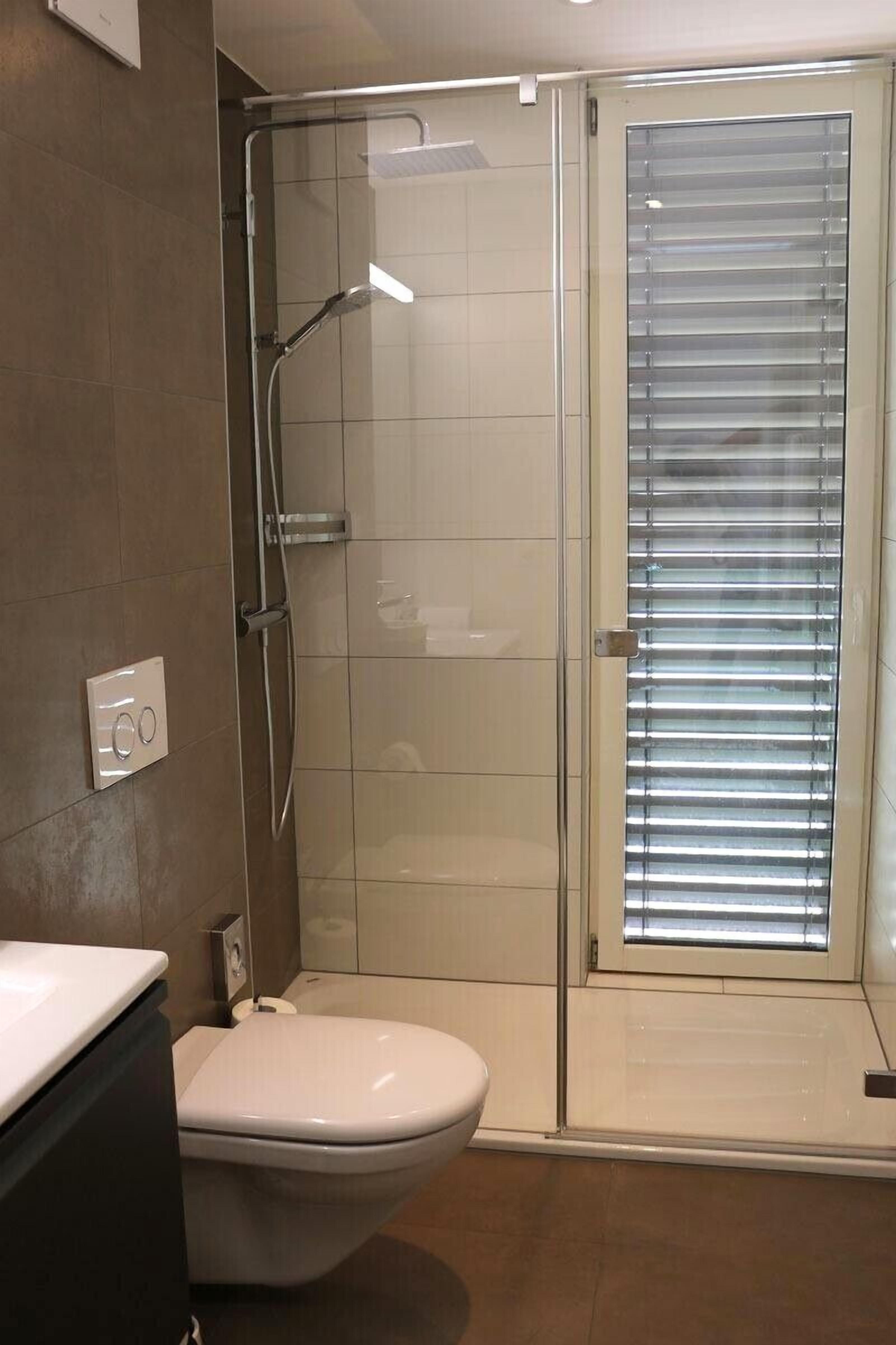 A modern bathroom with a glass shower enclosure, a toilet, and a window with blinds.