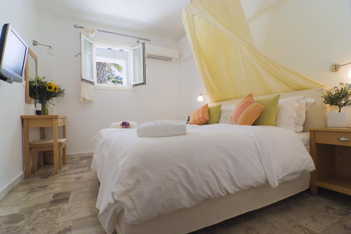 Un dormitorio ordenado con cama, mesita de noche, una pequeña silla de madera y televisor, además de una ventana con cortinas.