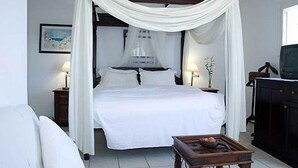 Un dormitorio con cama con dosel, ropa de cama blanca, dos mesitas de noche con lámparas, una mesita de noche de madera y una pequeña mesa de madera con fruta.