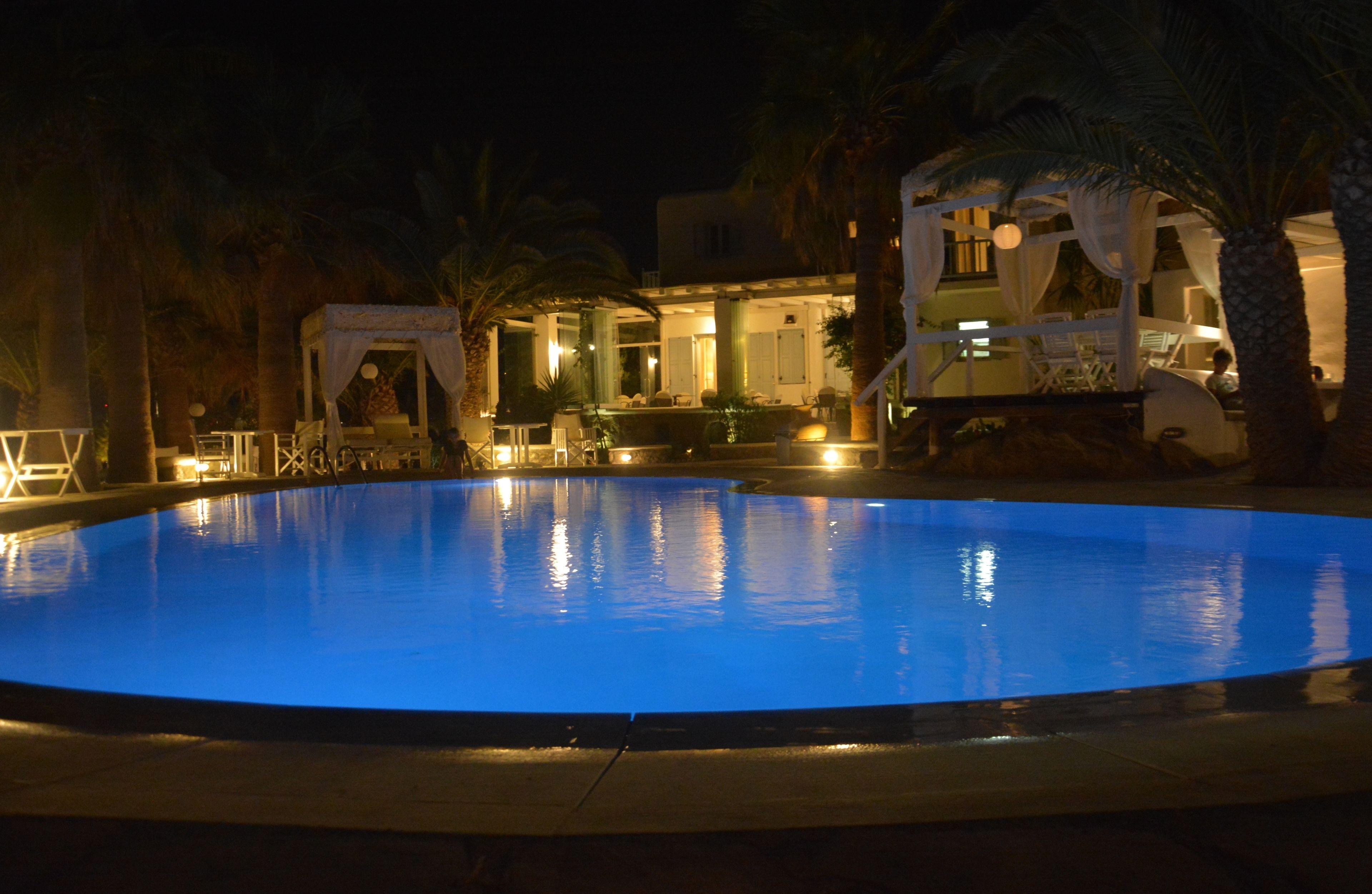 A nighttime pool area with a well-lit pool, lounge chairs, and a building with a covered patio.