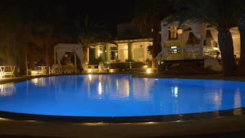 A nighttime pool area with a well-lit pool, lounge chairs, and a building with a covered patio.