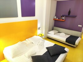 Une chambre d’hôtel avec un grand lit, un lit plus petit, une table de chevet avec une lampe, une étagère fixée au mur avec une bouteille, et une horloge murale.