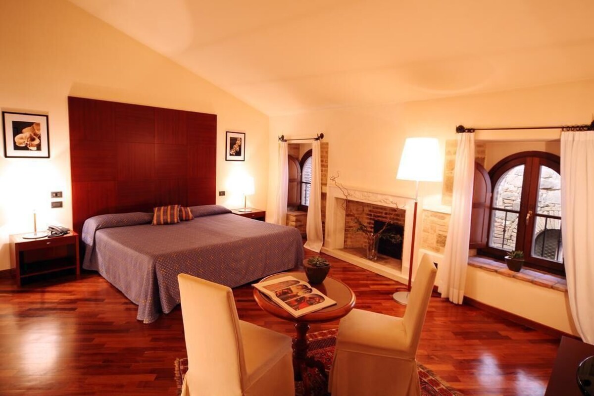 Une chambre à coucher avec un grand lit, une cheminée, une fenêtre avec des rideaux et une petite table sur laquelle se trouve un magazine.