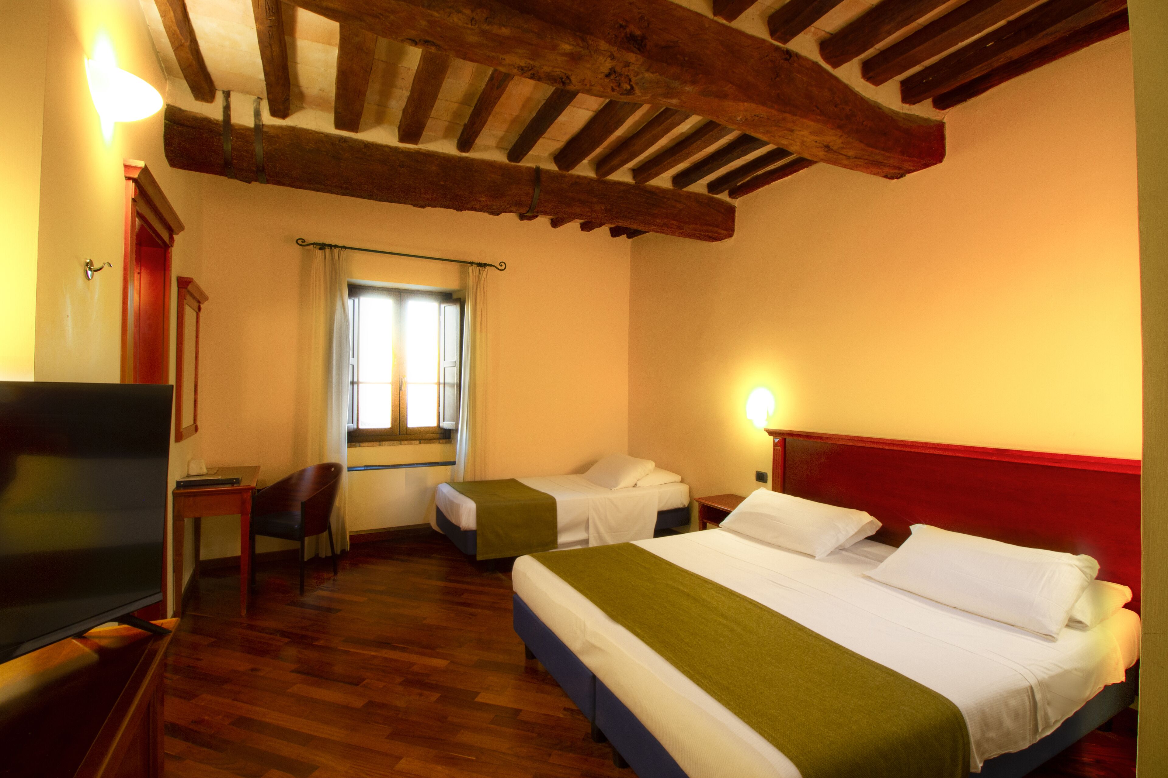A hotel room with wooden flooring, two beds with white linens, a television, a desk, and a window with curtains.