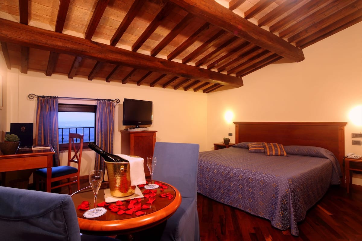 A cozy room with a wooden ceiling, a bed with patterned bedding, a TV on a wooden cabinet, a table with a bottle and glasses, and a view through a window with curtains.
