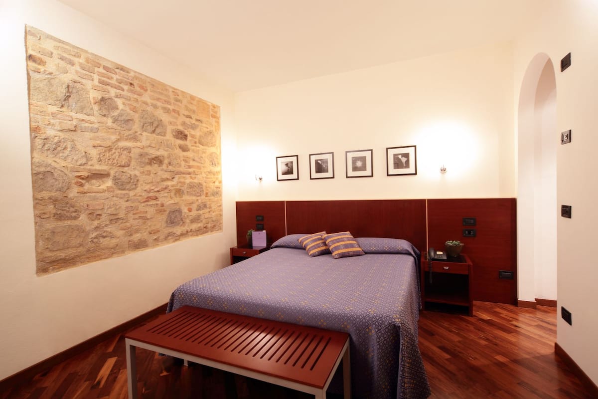 A bedroom with a stone wall, a bed with a headboard, bedside tables, a bench, and framed pictures on the wall.