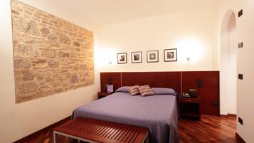 Un dormitorio con pared de piedra, cama con cabecera, mesitas de noche, un banco y cuadros enmarcados en la pared.