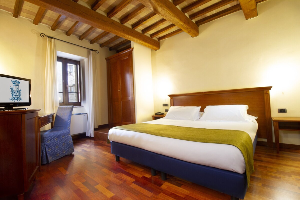 A hotel room with a wooden bed, a desk with a computer, a chair, and a window with curtains.