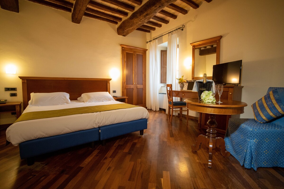 A hotel room with a double bed, wooden flooring, a small table with a vase and glass, a chair, and a TV.
