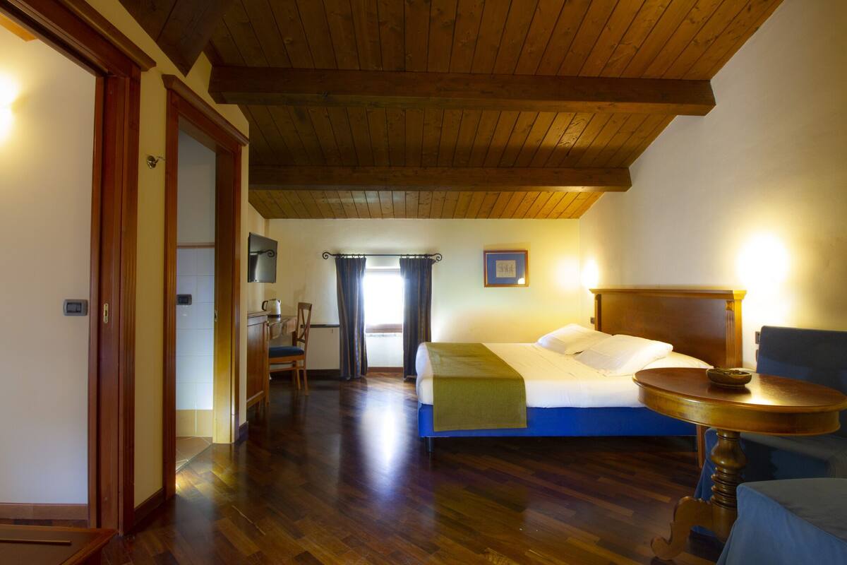 A bedroom with a wooden ceiling, a bed with a blue headboard, a bedside table, and a chair.