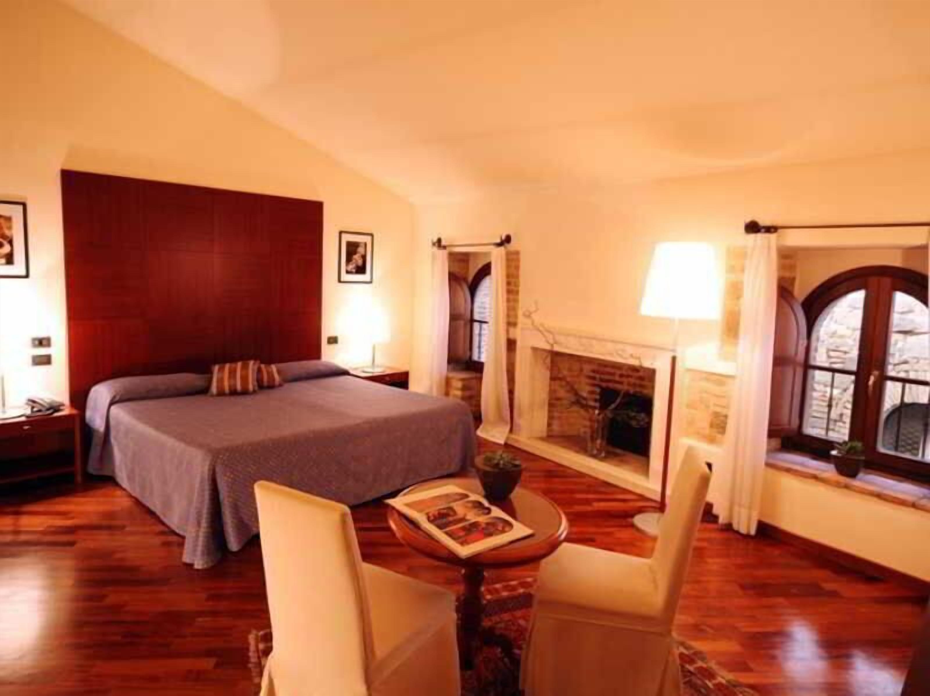 Una camera d'albergo con un letto grande, un tavolino con un libro, due poltrone, un camino e una finestra con tende.
