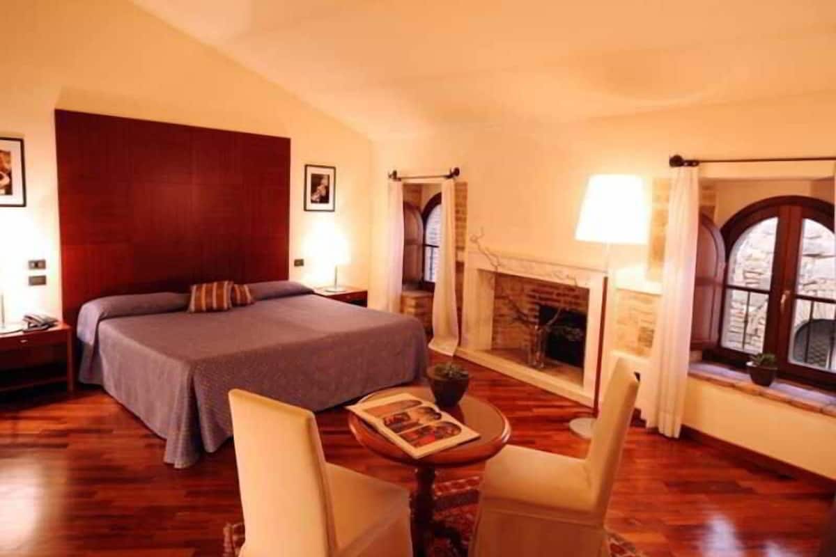 A hotel room with a large bed, a small table with a book, two chairs, a fireplace, and a window with curtains.
