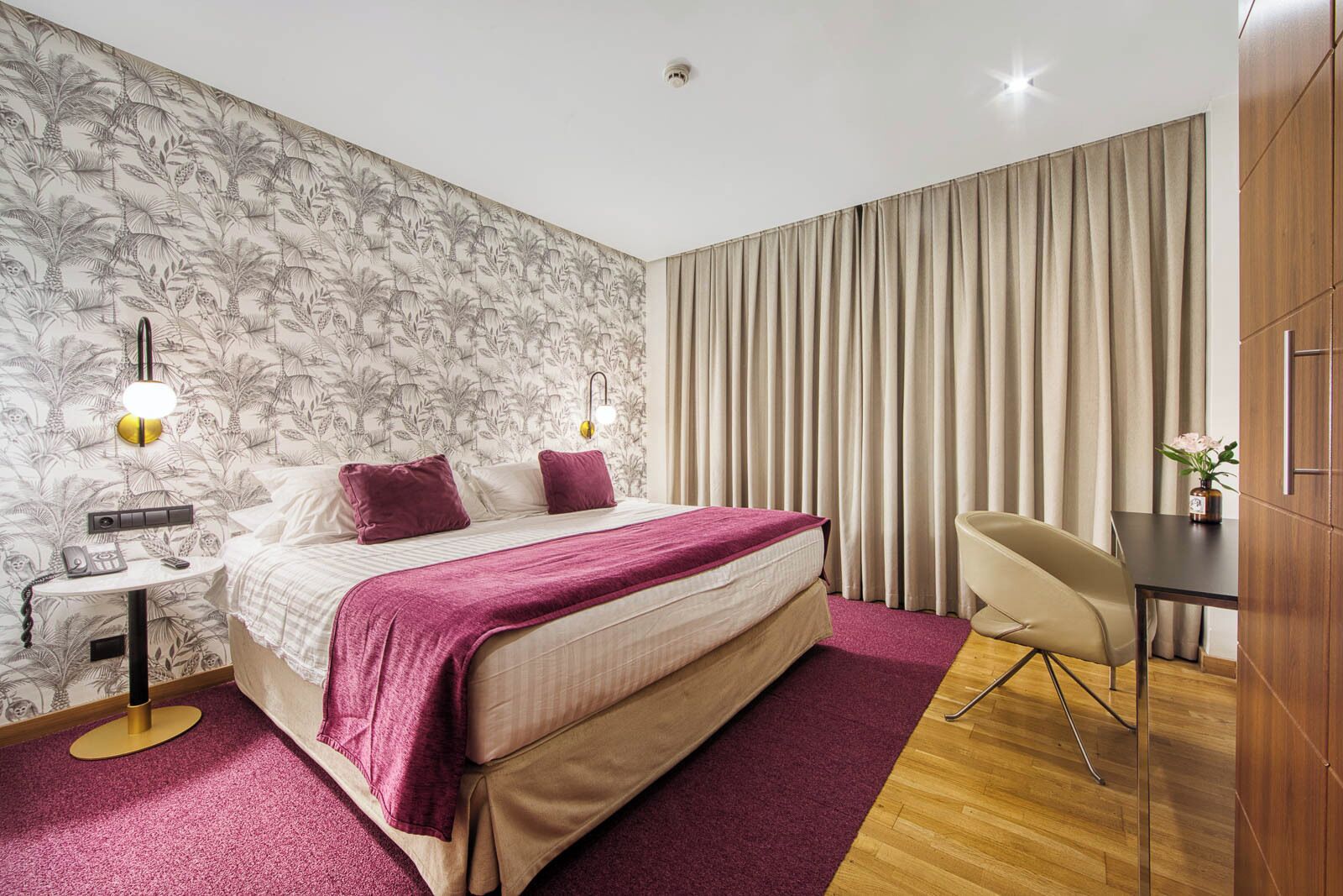 A hotel room with a bed, a desk, a chair, a nightstand, and a wall with a tropical wallpaper.