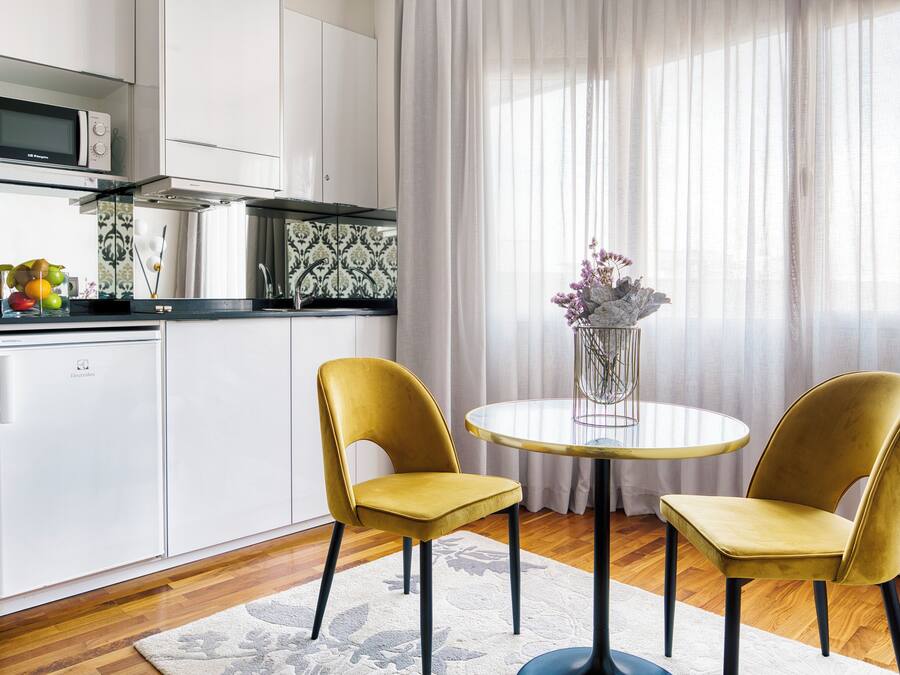 Una cocina moderna con armarios blancos, un microondas y un frutero. Hay un comedor con dos sillas amarillas y una mesa redonda pequeña.