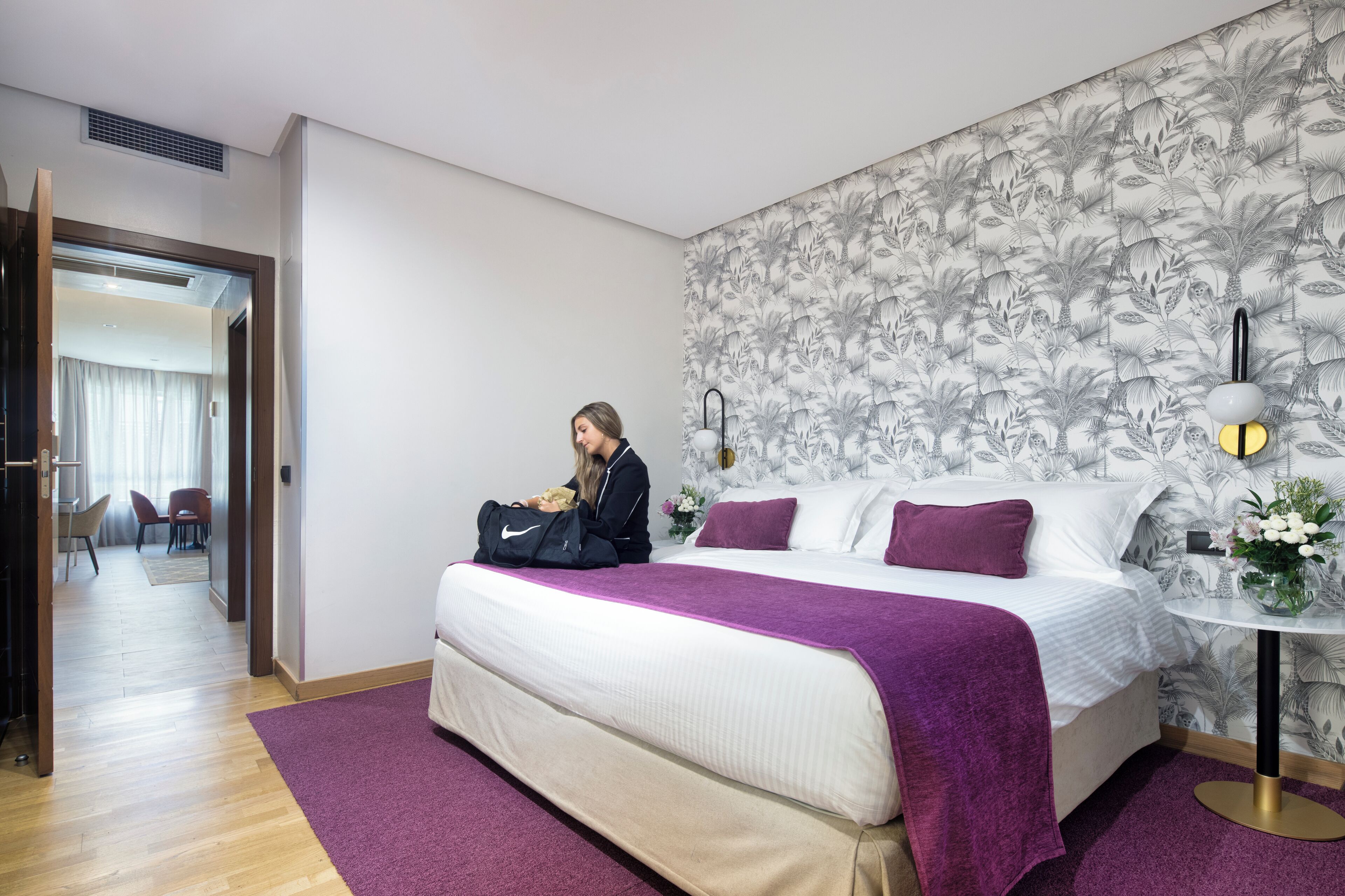 Habitación de hotel con cama, una manta morada, una mesita de noche con lámpara y papel tapiz floral.