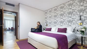 A hotel room with a bed, a purple throw, a nightstand with a lamp, and a floral wallpaper.
