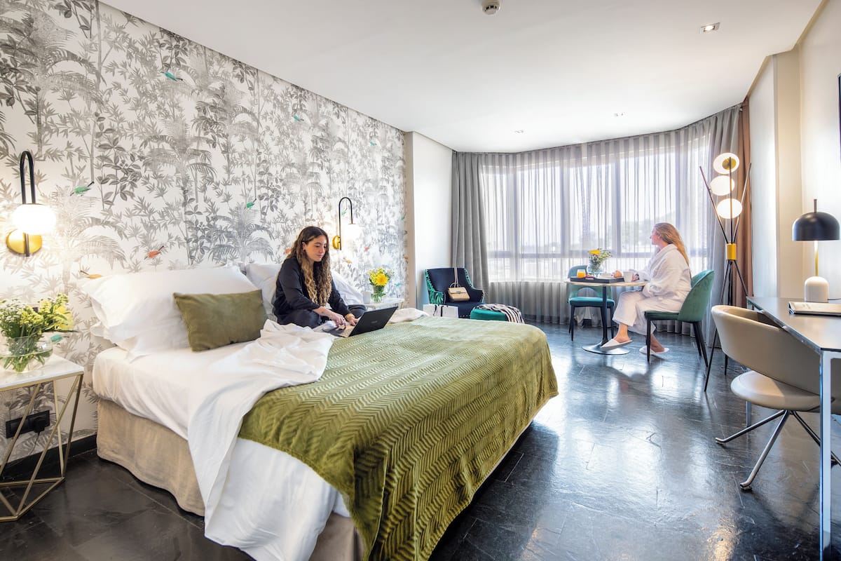 Een hotelkamer met een bed, een bureau en een stoel. Er is een groot raam met gordijnen.