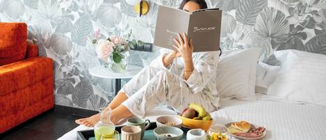 Uma pessoa lendo um livro na cama, com uma bandeja de itens de café da manhã, incluindo frutas, uma caneca e um copo de suco de laranja.