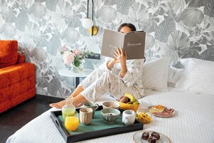 Una persona leyendo un libro en una cama con una bandeja de desayuno que incluye frutas, una taza y un vaso de zumo de naranja.