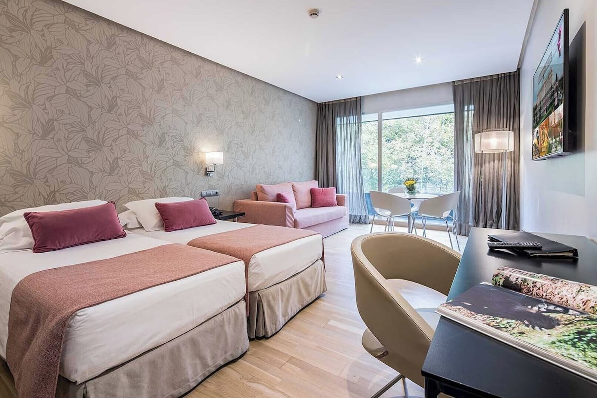 Hotelkamer met twee bedden, een bureau en een stoel. Er is een raam met gordijnen, een aan de muur bevestigde televisie en behangpapier met een bloemmotief.