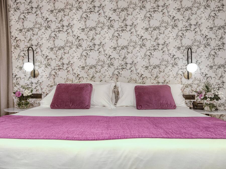 Quarto de hotel com papel de parede floral, duas almofadas roxas, um cobertor rosa e mesas de cabeceira com abajures.