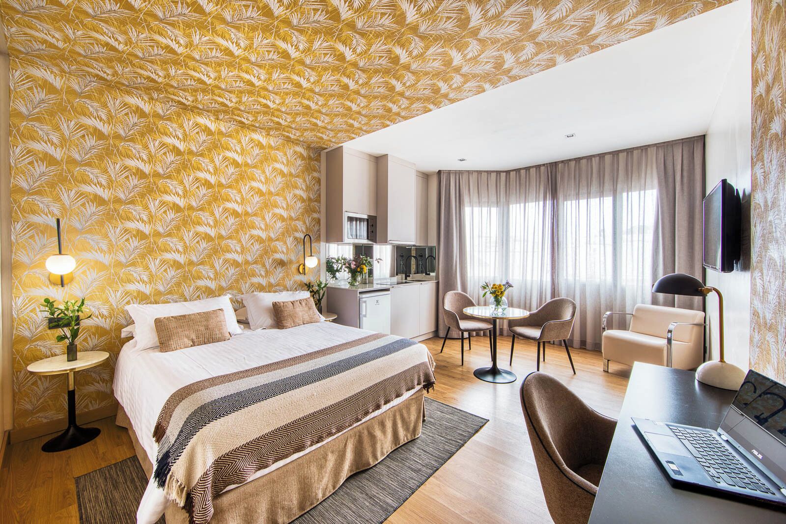 Quarto de hotel com cama, escrivaninha com laptop, área de refeições com cadeiras, televisão e cozinha.