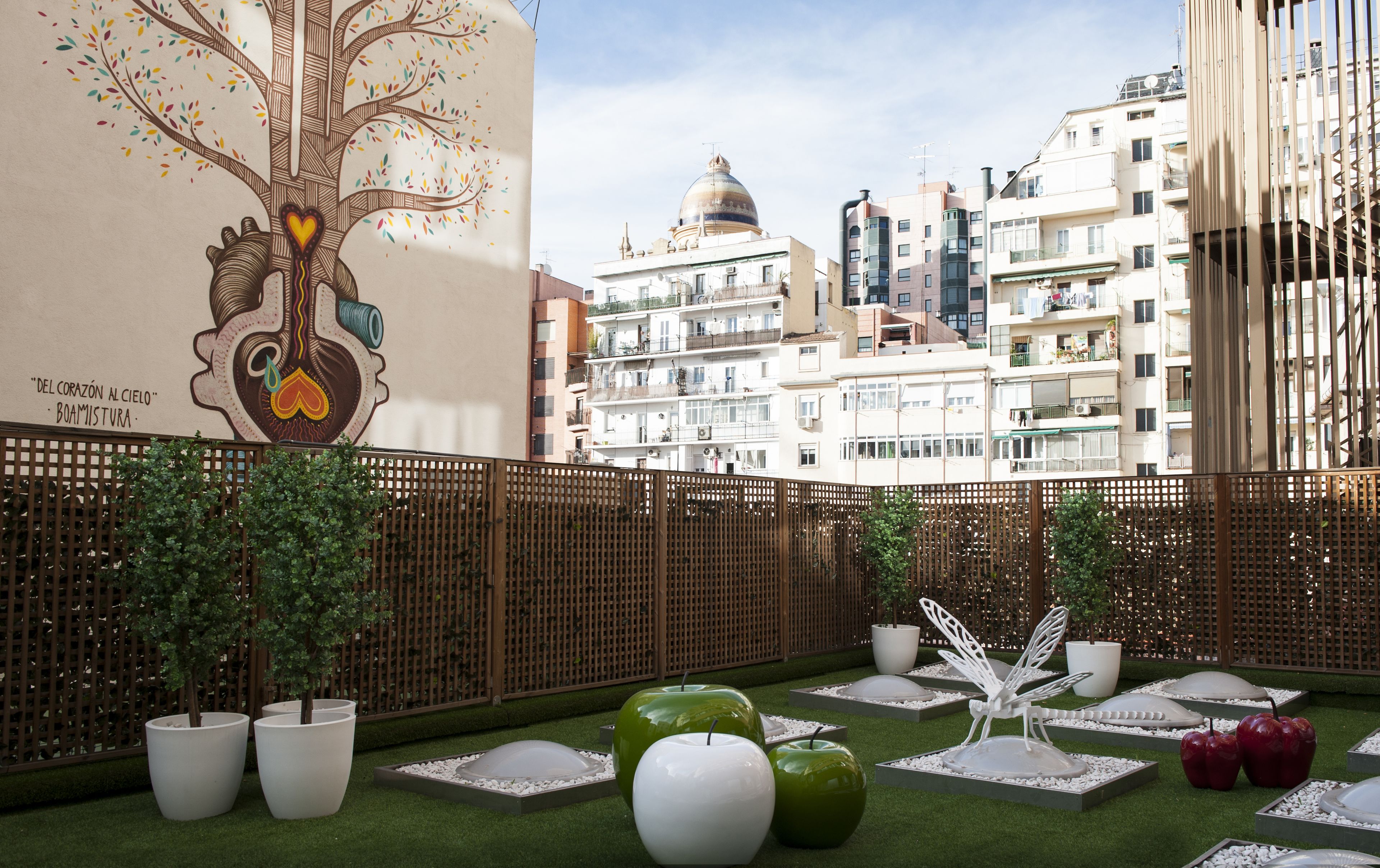 A rooftop garden with potted plants, a decorative tree mural, and a view of city buildings.
