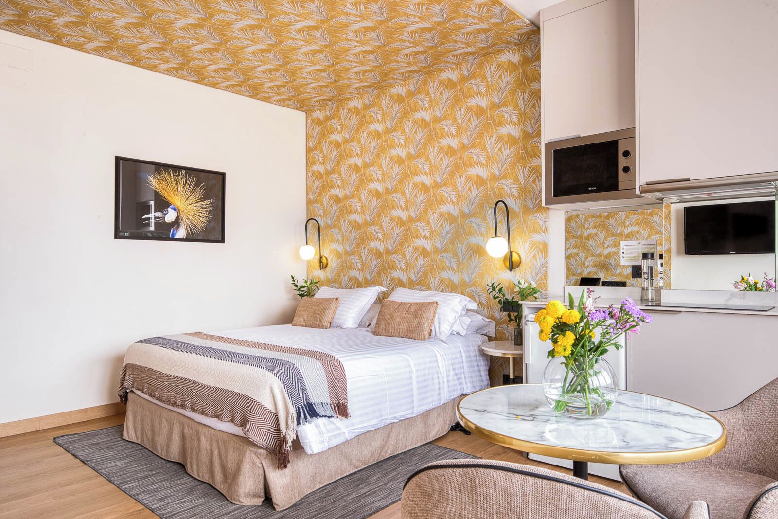 Habitació d'hotel moderna amb un llit, una taula de nit, una taula rodona petita amb un ram de flors, un sofà i una zona de cuina equipada.