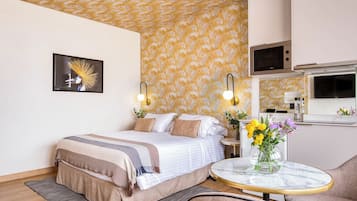 Habitació d'hotel moderna amb un llit, una taula de nit, una taula rodona petita amb un ram de flors, un sofà i una zona de cuina equipada.