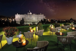 Un área de estar al aire libre con mesas y sillas, un edificio grande al fondo y un cielo nocturno lleno de estrellas.