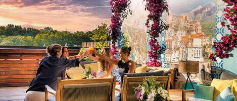 A terrace with wooden furniture, a table with a vase of flowers, and a mural of a coastal town.