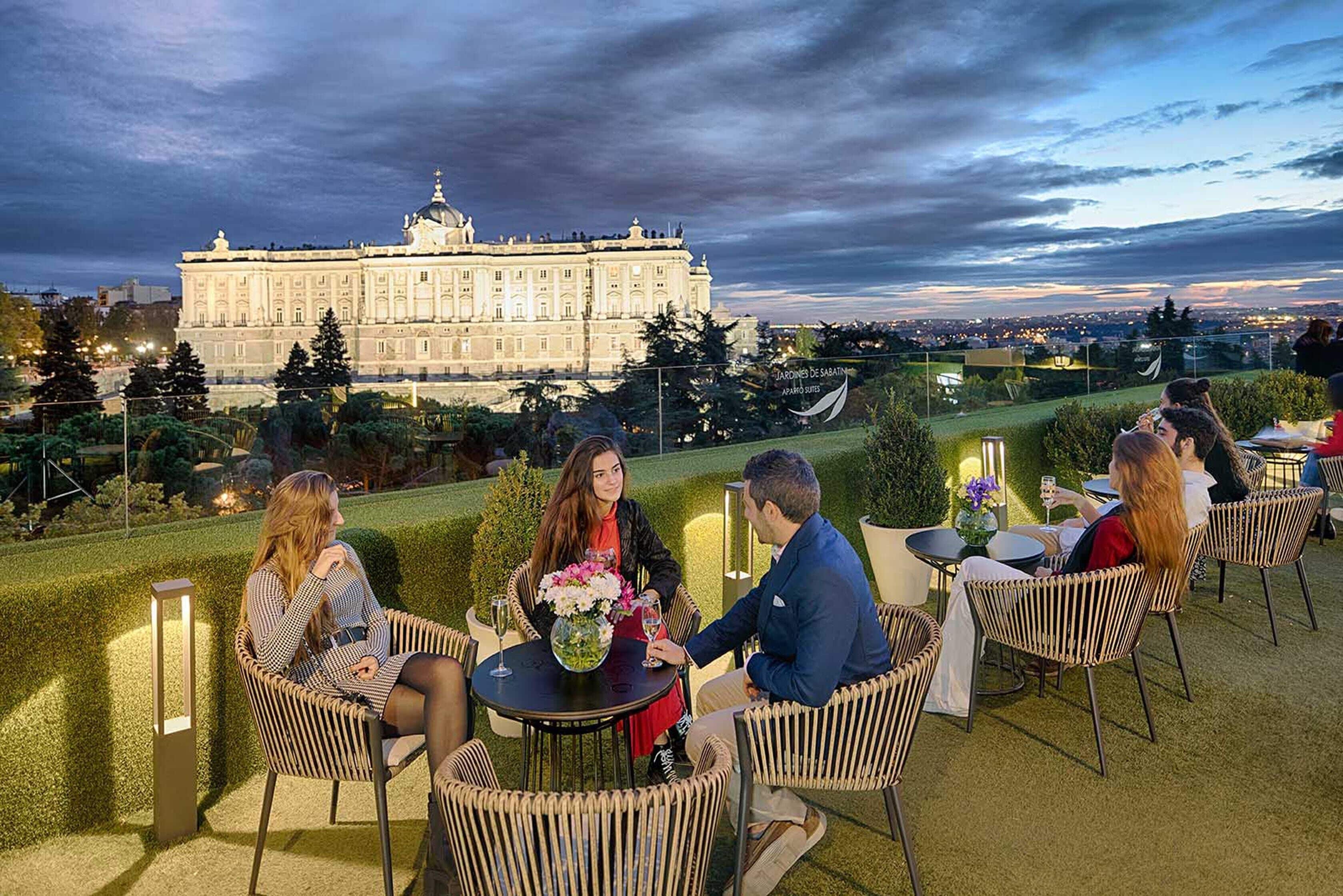 Un grupo de personas sentadas en mesas en una terraza al aire libre con vista a un edificio grande.