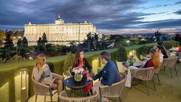 Un grupo de personas sentadas en mesas en una terraza al aire libre con vista a un edificio grande.