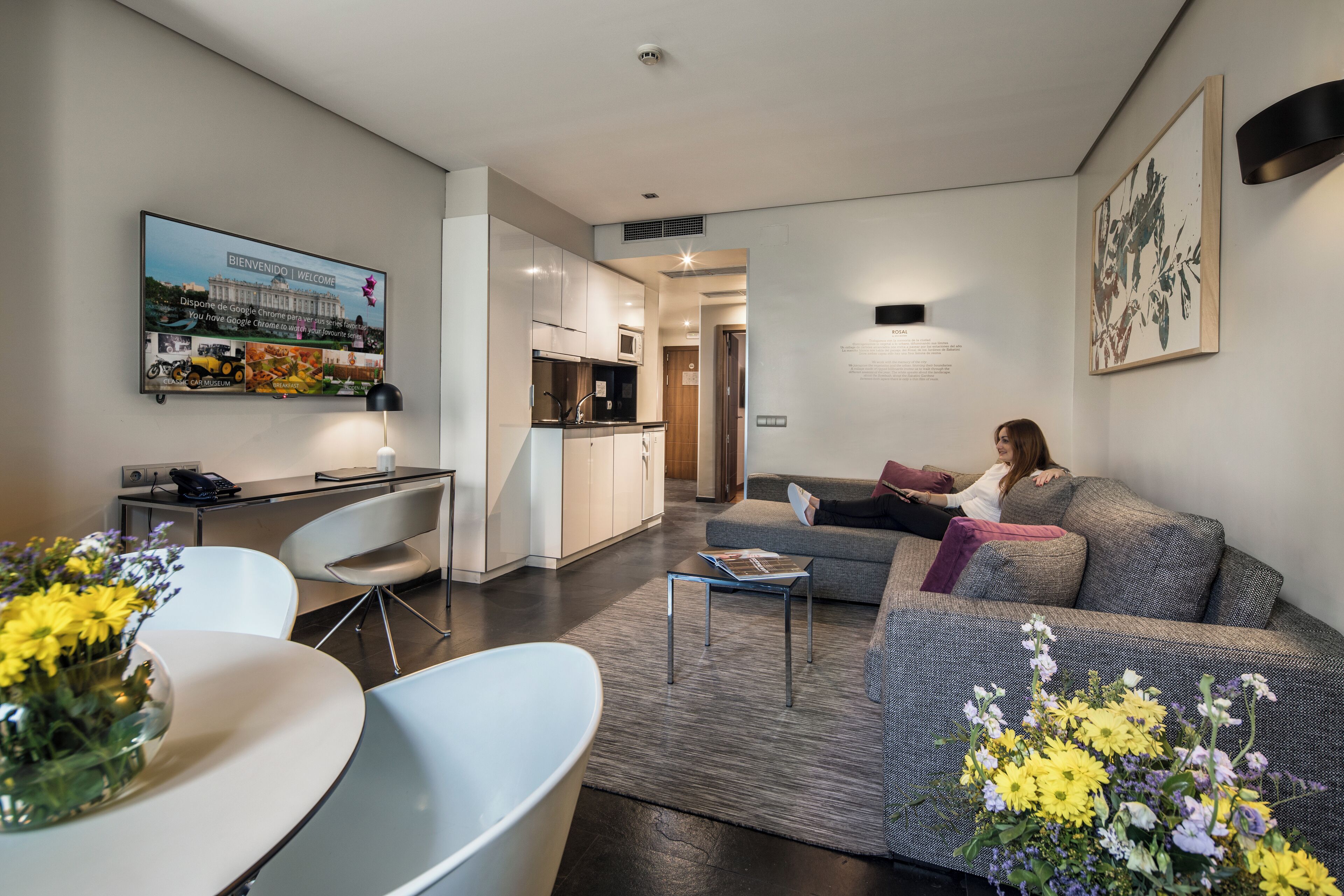 Een moderne woonkamer met een bank, salontafel en een eethoek met tafel en stoelen. Achtergrond toont een keuken met kastjes en een magnetron.