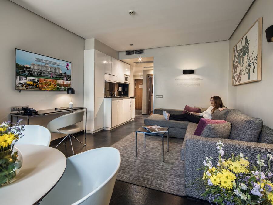 Een moderne woonkamer met een bank, salontafel en een eethoek met tafel en stoelen. Achtergrond toont een keuken met kastjes en een magnetron.