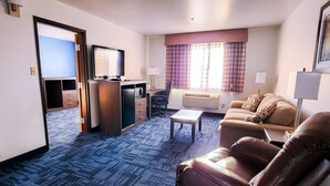 A hotel room with a blue carpet, a sofa, a chair, a small table, a TV, and a window with curtains.