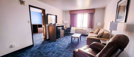 A hotel room with a blue carpet, a sofa, a chair, a small table, a TV, and a window with curtains.