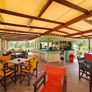 An outdoor cafe with wooden tables and chairs, a bar area, and a view of greenery. - Hotel Alex (Karpathos)