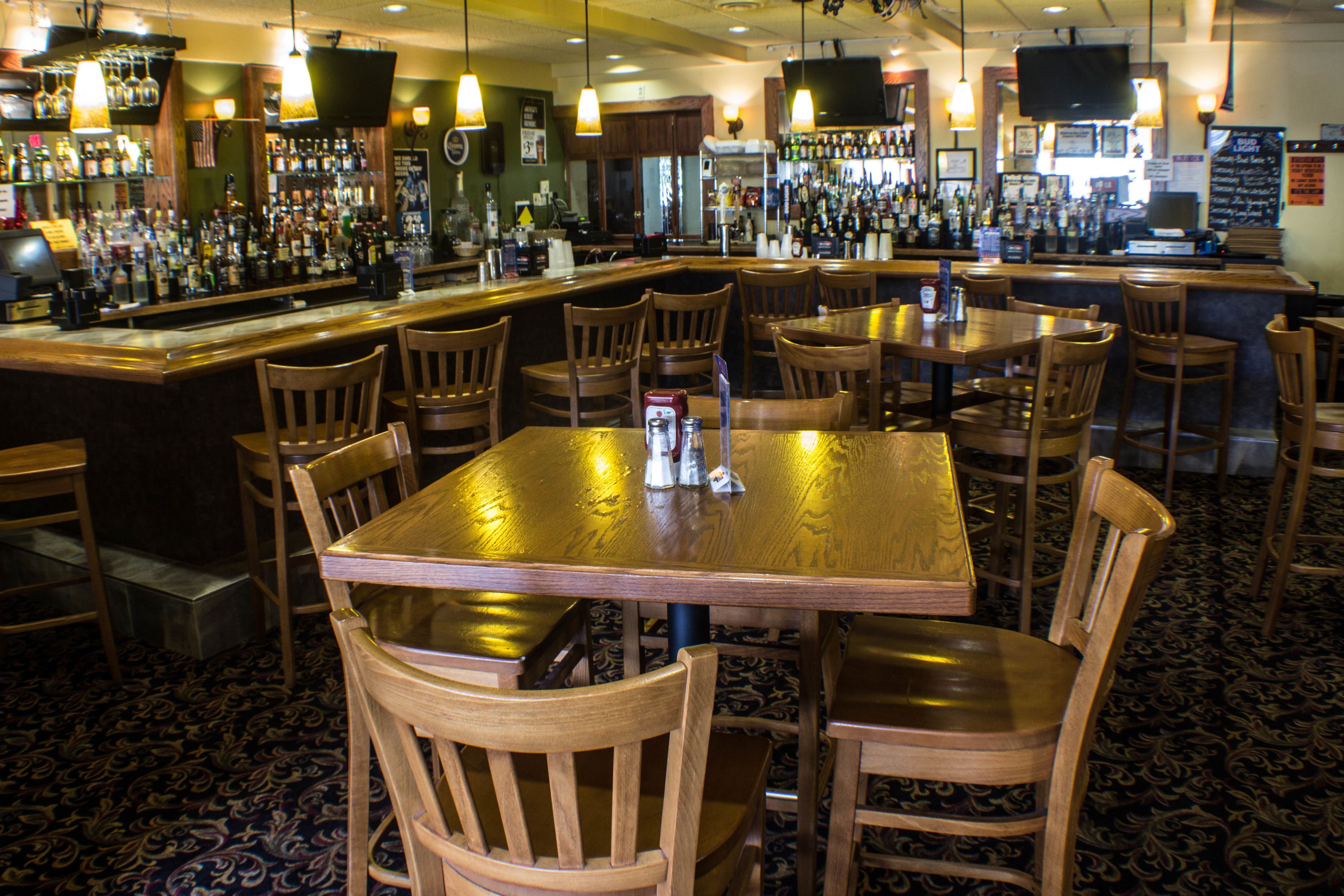 A bar area with wooden tables and chairs, a bar counter stocked with bottles, and a patterned carpet.
