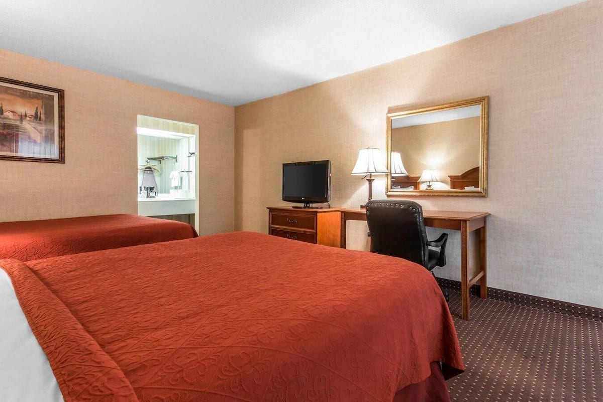 A hotel room with two beds, a desk with a television, a mirror, and a window with a view of the outside.