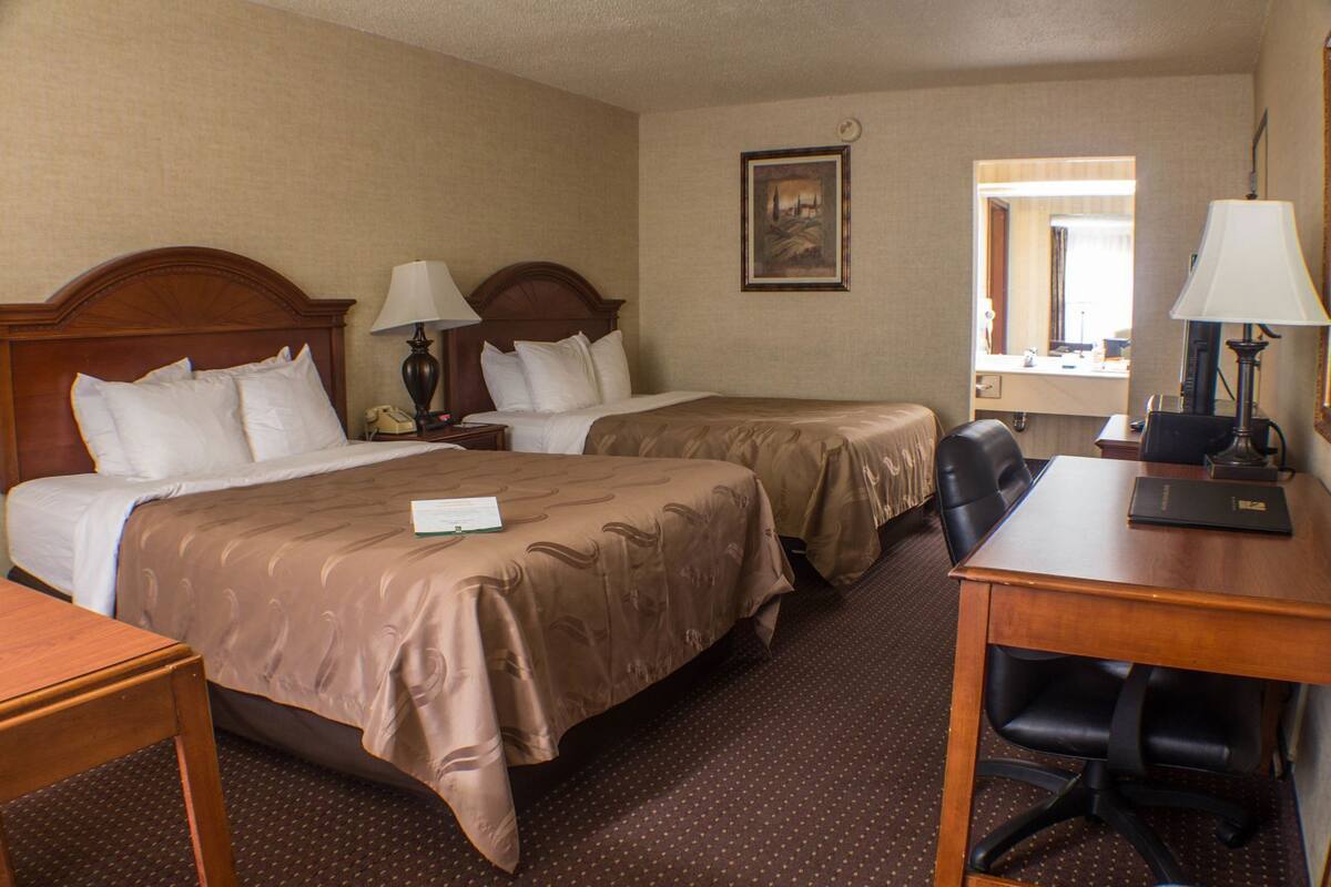 A hotel room with two beds, a desk, a chair, a lamp, a telephone, and a painting on the wall.
