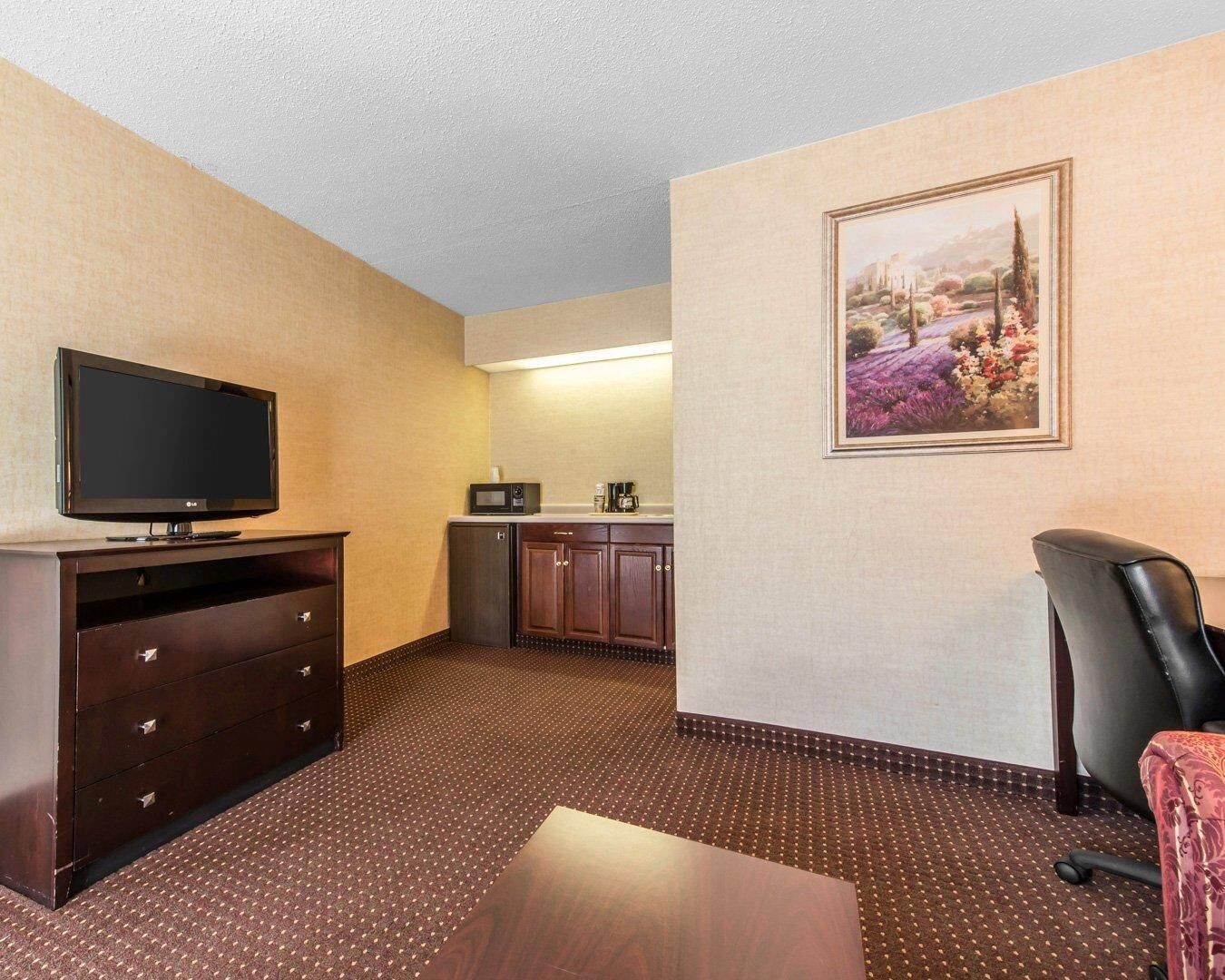 A hotel room with a flat-screen TV, a microwave, a small refrigerator, a dining table, a chair, and a framed landscape painting on the wall.