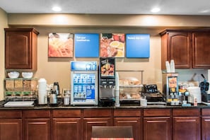 A hotel breakfast area with a self-serve station, coffee machines, and a refrigerated display case.