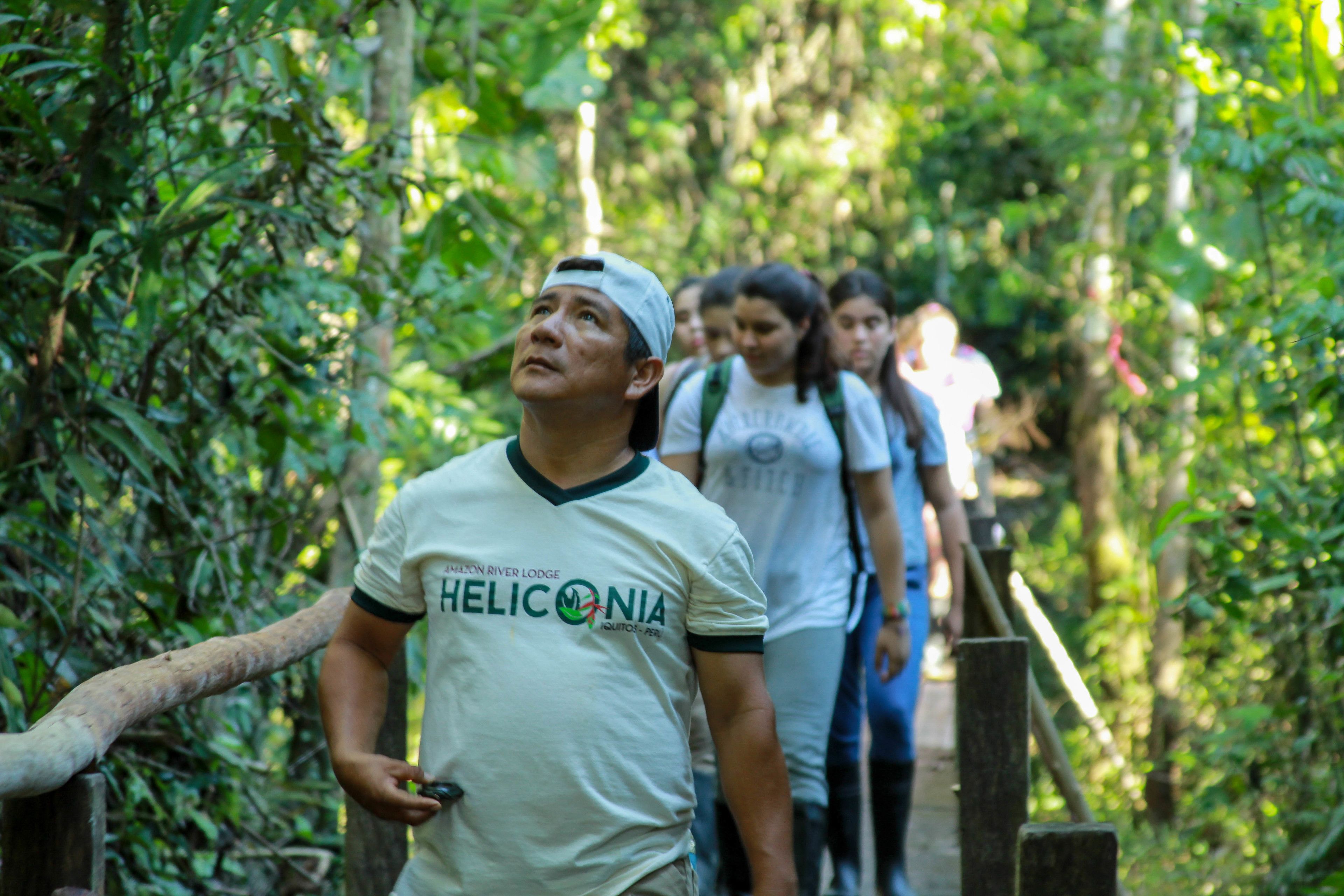 A person wearing a white t-shirt with a logo, walking on a wooden bridge surrounded by greenery.