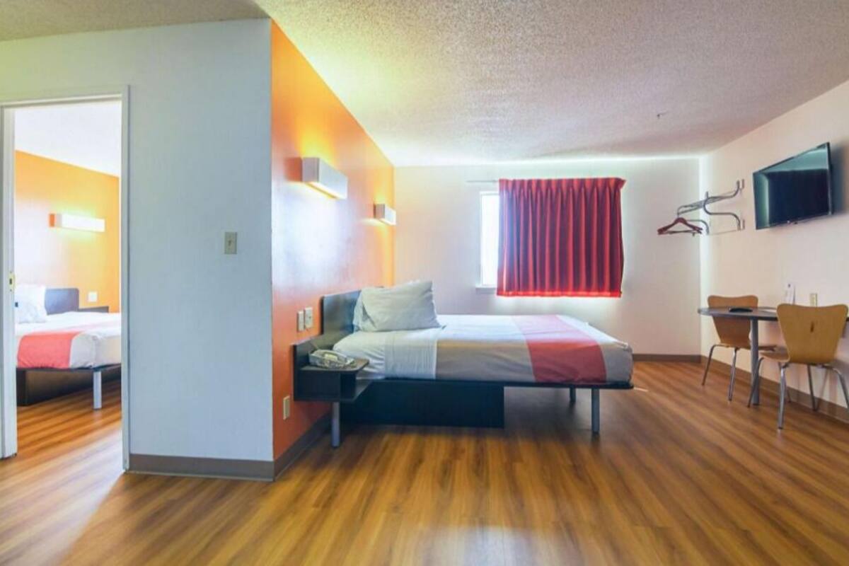 A modern hotel room with a wooden floor, a bed with a red and white bedspread, a desk with a chair, and a TV mounted on the wall.