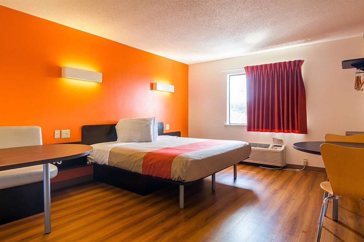 A hotel room with a bed, a desk, a chair, a window with red curtains, and an air conditioning unit.