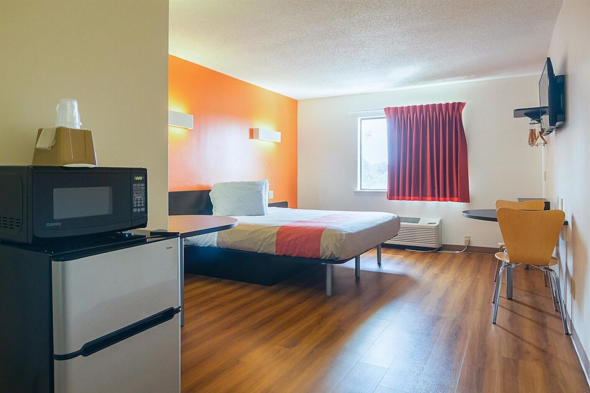 A compact hotel room with a bed, a desk, a chair, a microwave, and a heater.