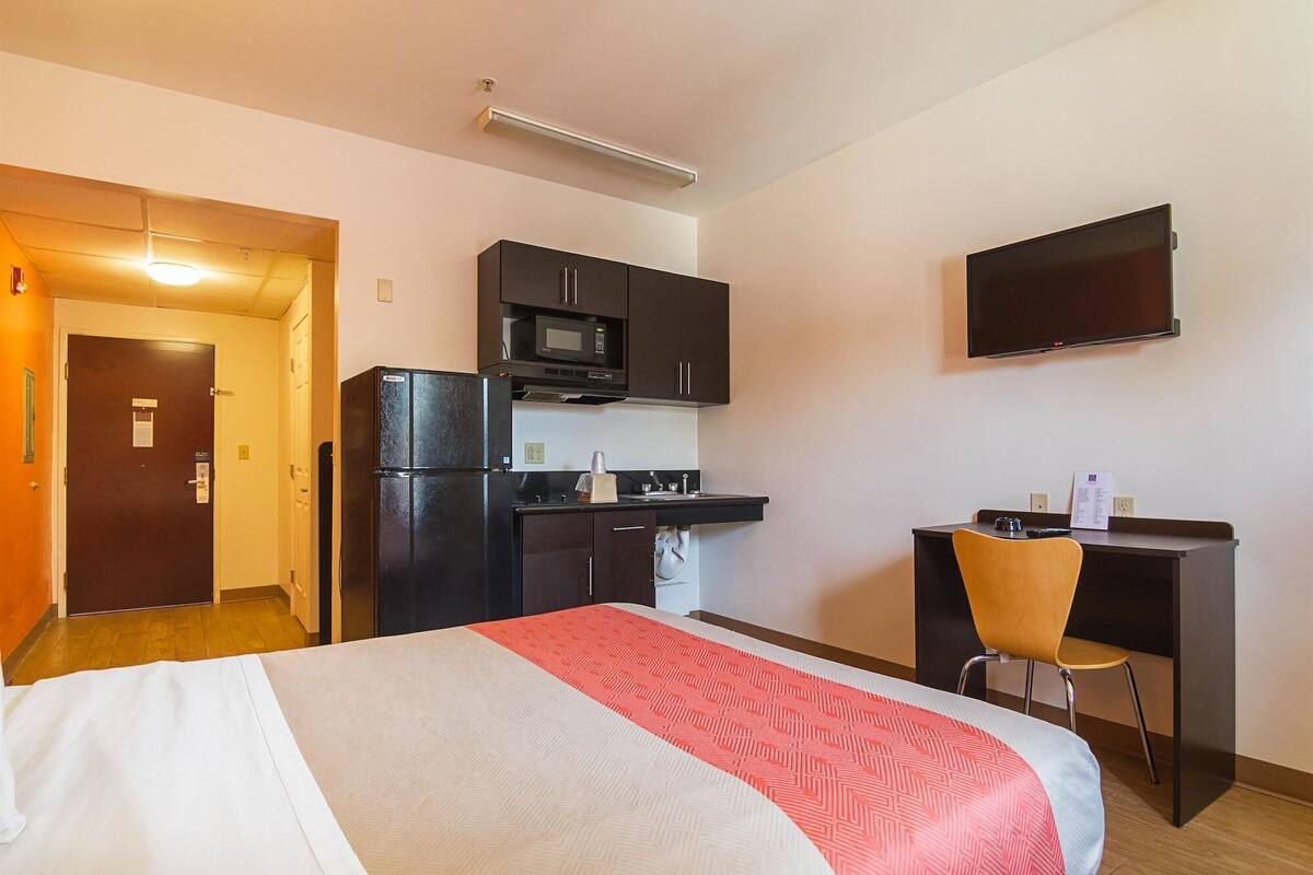 A compact hotel room with a bed, kitchenette, and a desk with a chair.