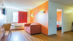 A modern hotel room with a wooden floor, a red sofa, a dining area with chairs, and a window with red curtains. - Motel 6 Jonesboro, GA (Jonesboro)