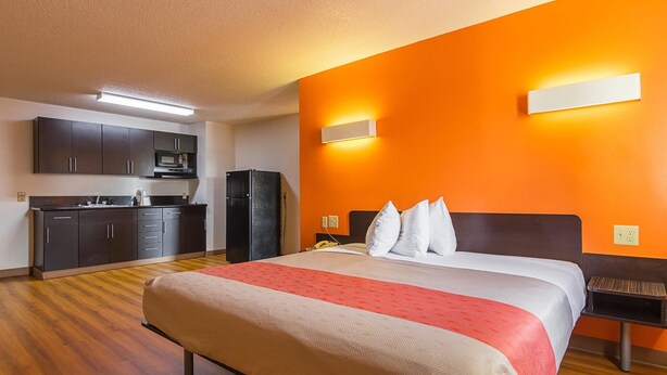 A hotel room with a bed, a bench, a kitchenette, and an orange accent wall. - Motel 6 Jonesboro, GA (Jonesboro)